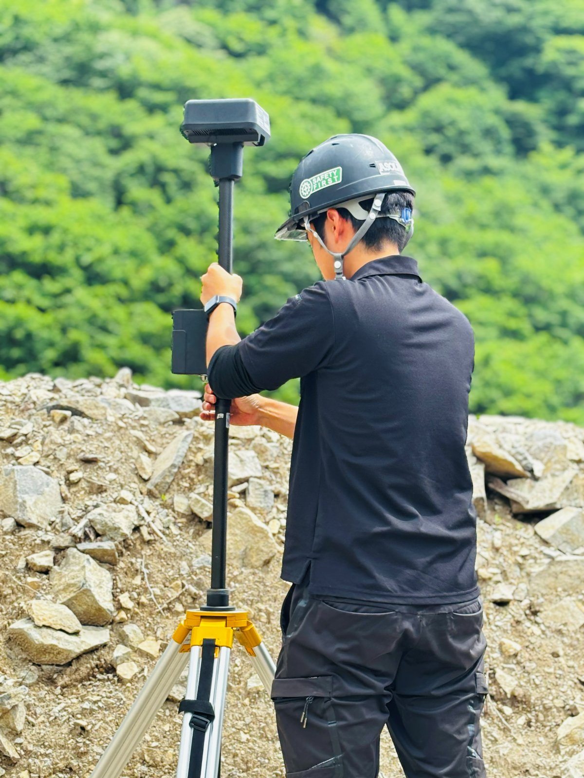 ヘルメット姿の作業員が、上端に黒い箱型のGNSSアンテナを付けた測量ポールを支えている後ろ姿。背景は採石場の岩と山の樹林