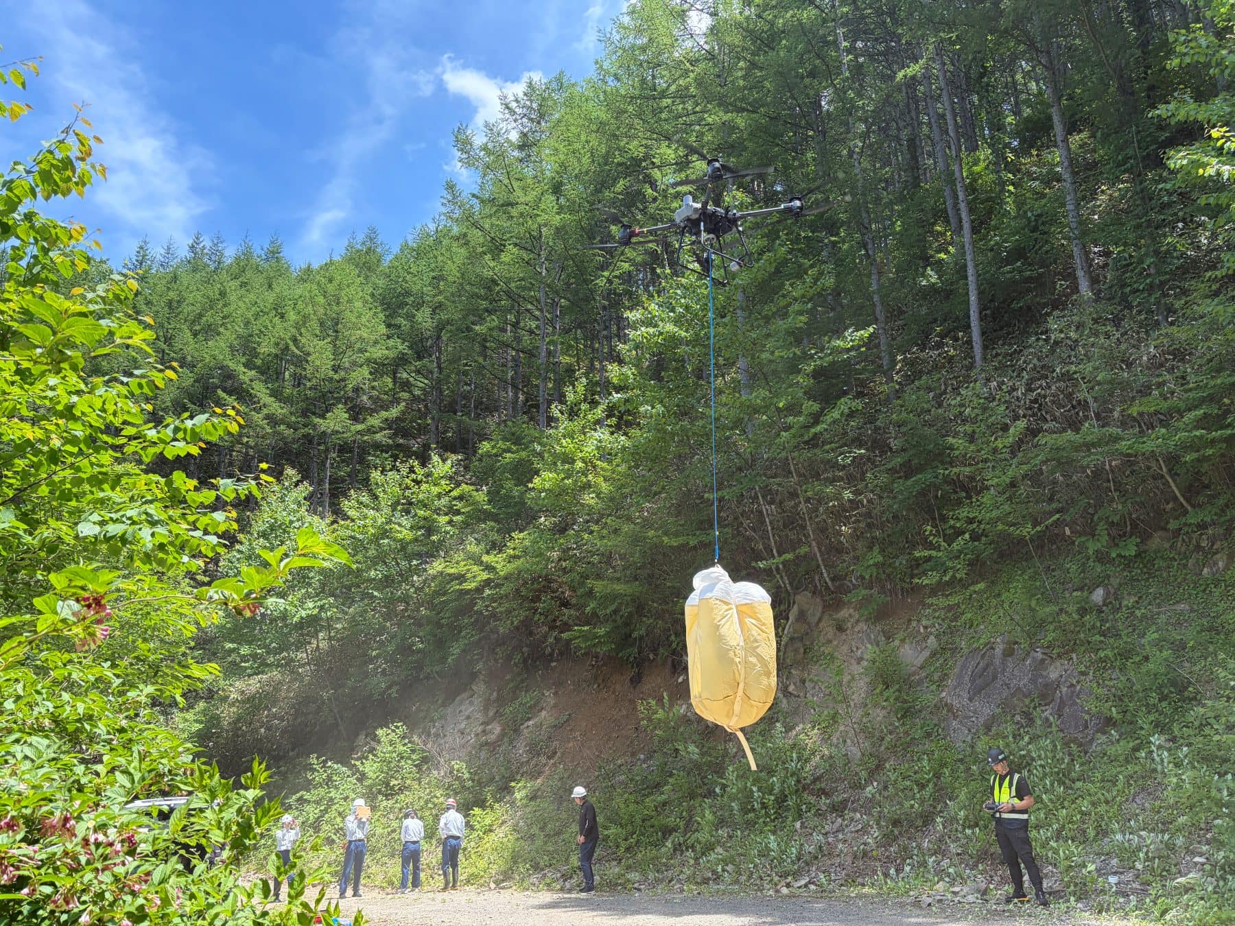 苗木を入れた黄色いフレコンバッグを吊り下げて、林道の上を飛ぶ DJI FlyCart 30。林道で作業員が見守っている
