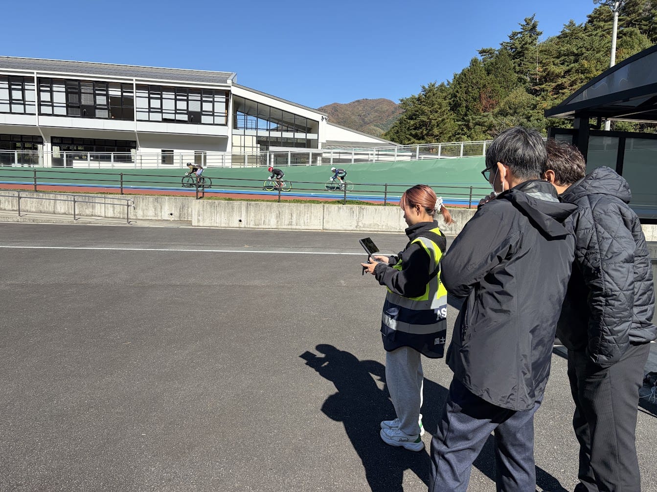 送信機の画面で撮影中の映像を確認する当社のドローン操縦者と、松本市の職員